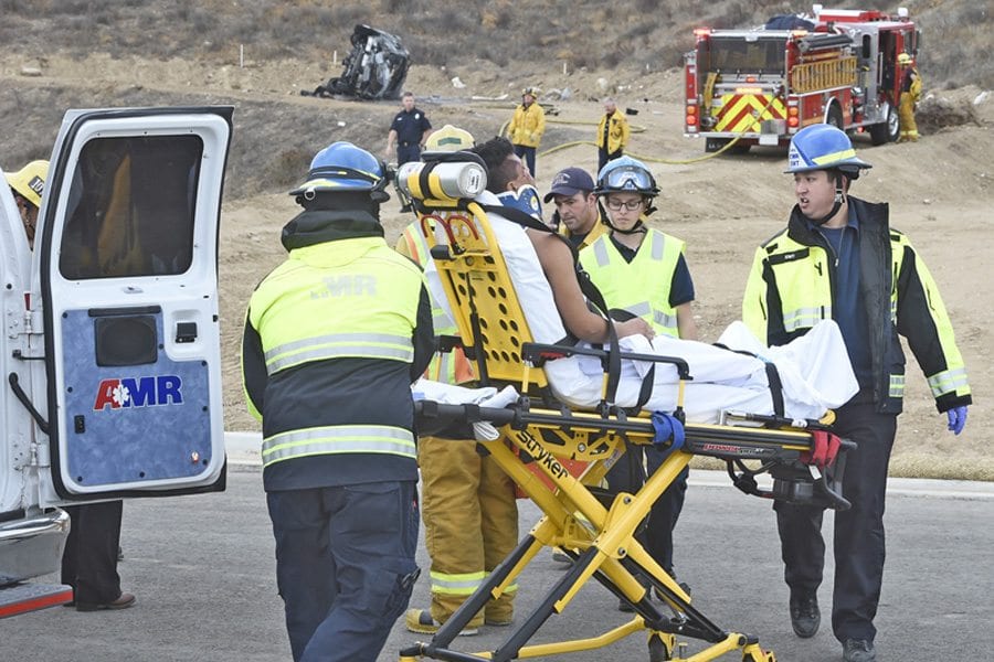 The occupant of a vehicle that rolled down a hill and caught fire (seen in the distance) is transported by ambulance from the Plum Canyon area on Saturday. Dan Watson/The Signal
